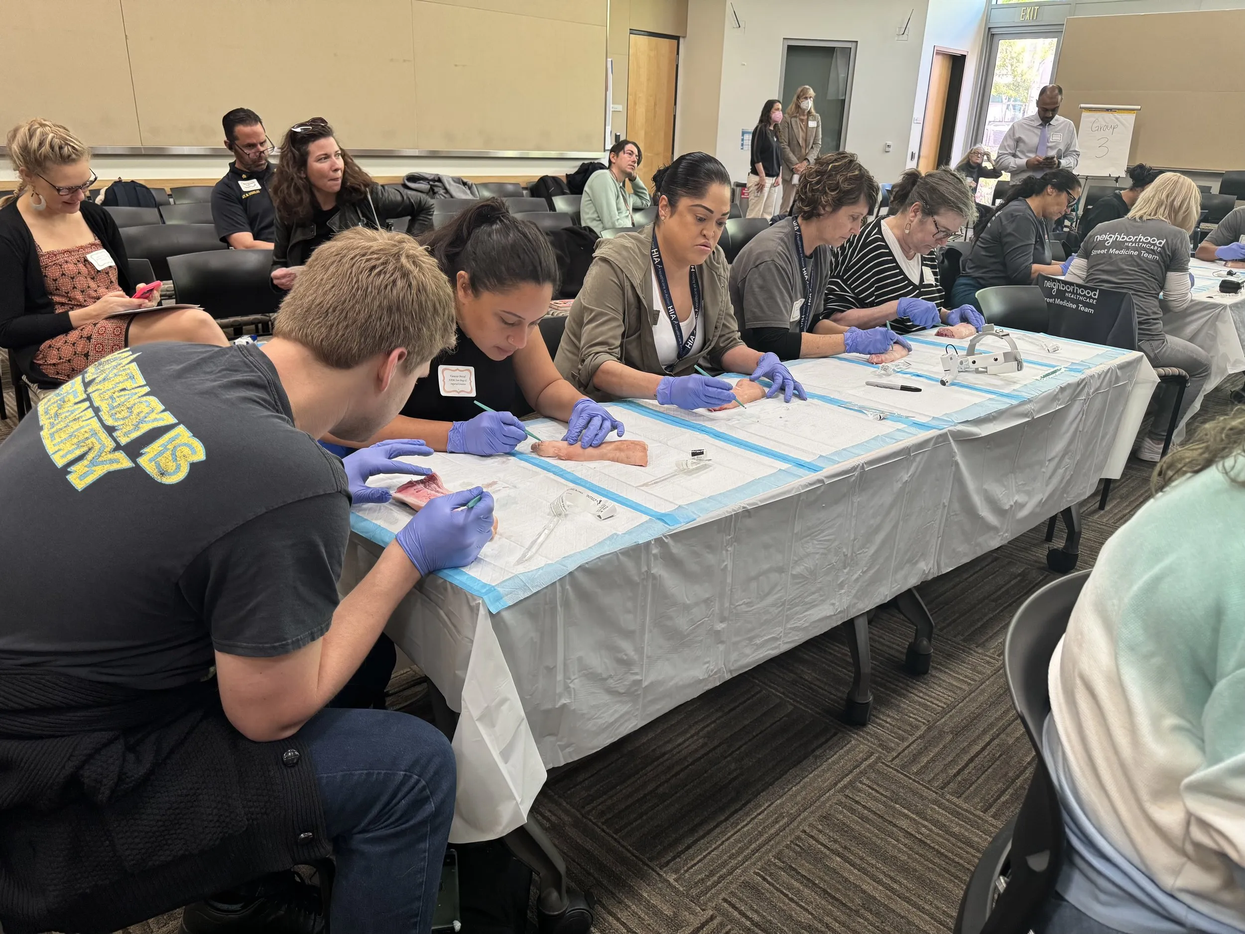 People sitting at tables in a classroom setting, participating in a hands-on training session. Some individuals wear gloves and engage with materials or tools on the table.