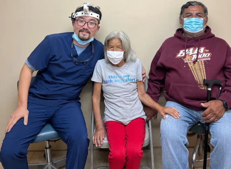 A healthcare professional in blue scrubs and two elderly individuals sitting together, all wearing face masks.