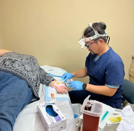 A medical professional in scrubs and magnifying glasses performs a procedure on a patient's hand in a clinic.