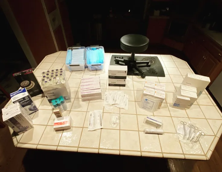 Medical supplies on a tiled kitchen counter including gloves, syringes, vials, and surgical instruments.