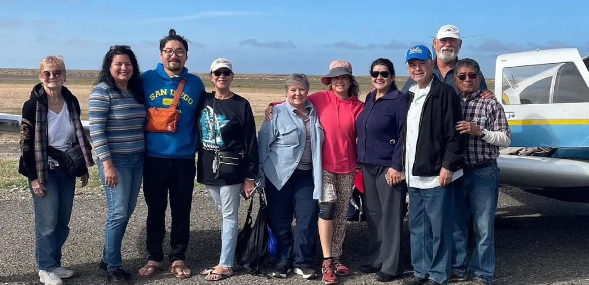 Group of people posing outdoors by a small plane on a sunny day.