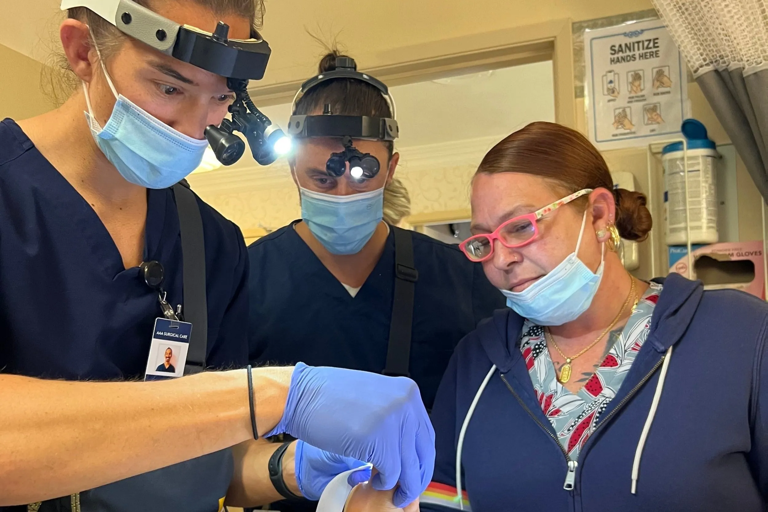 Medical professionals in masks and scrubs, performing a procedure with surgical tools in a clinical setting.