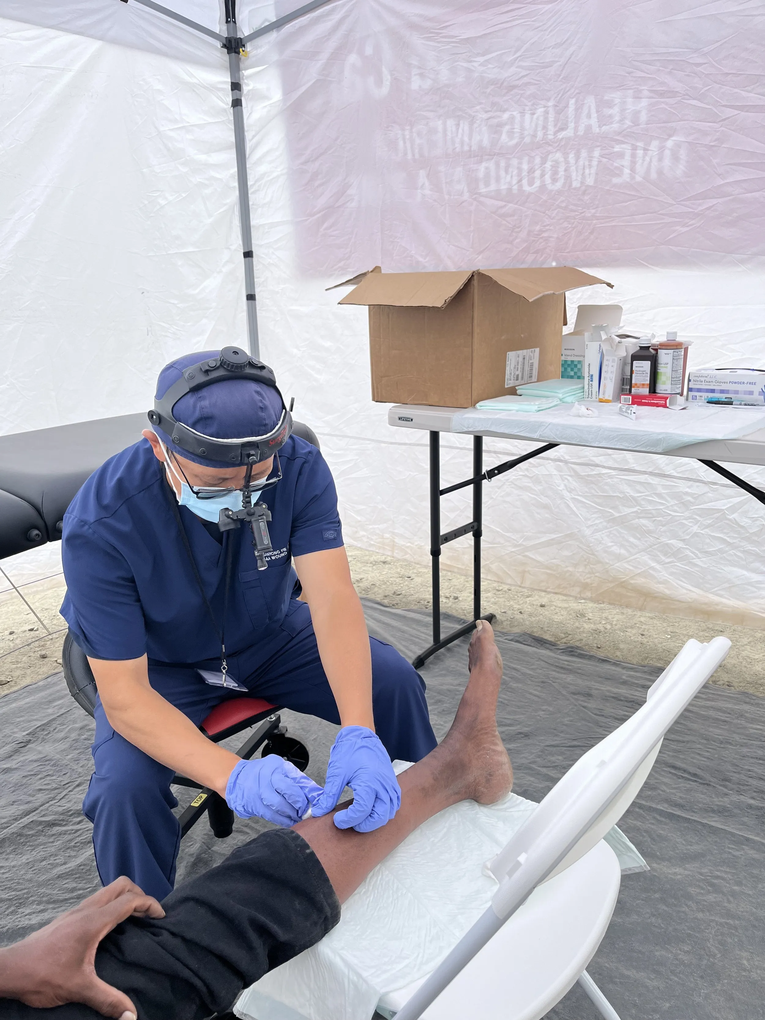 Medical professional in blue scrubs and gloves treating a patient's leg in a mobile medical tent, with supplies on a nearby table.