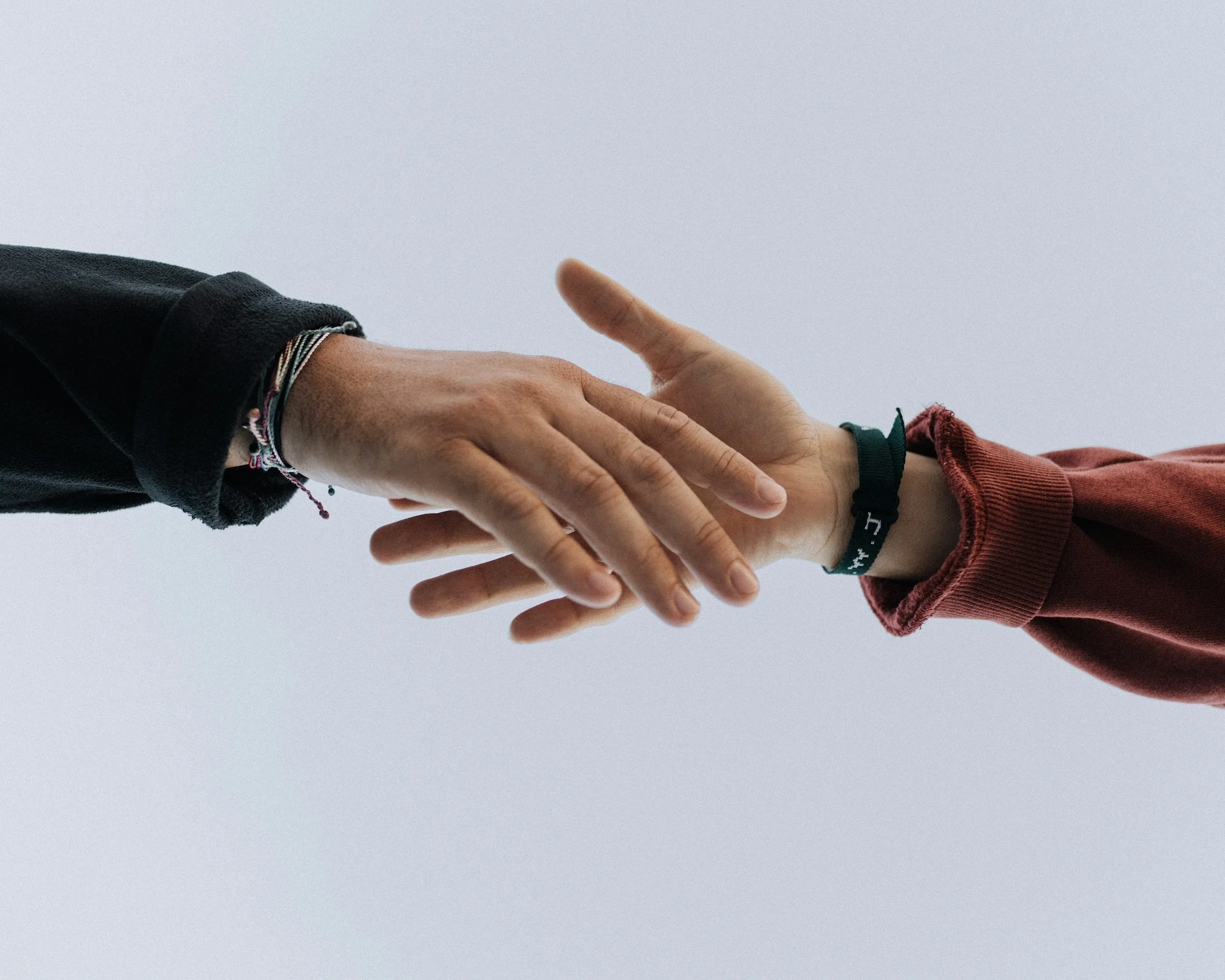 Two hands reaching out toward each other against a light blue background, with one wearing bracelets and the other a green wristband.