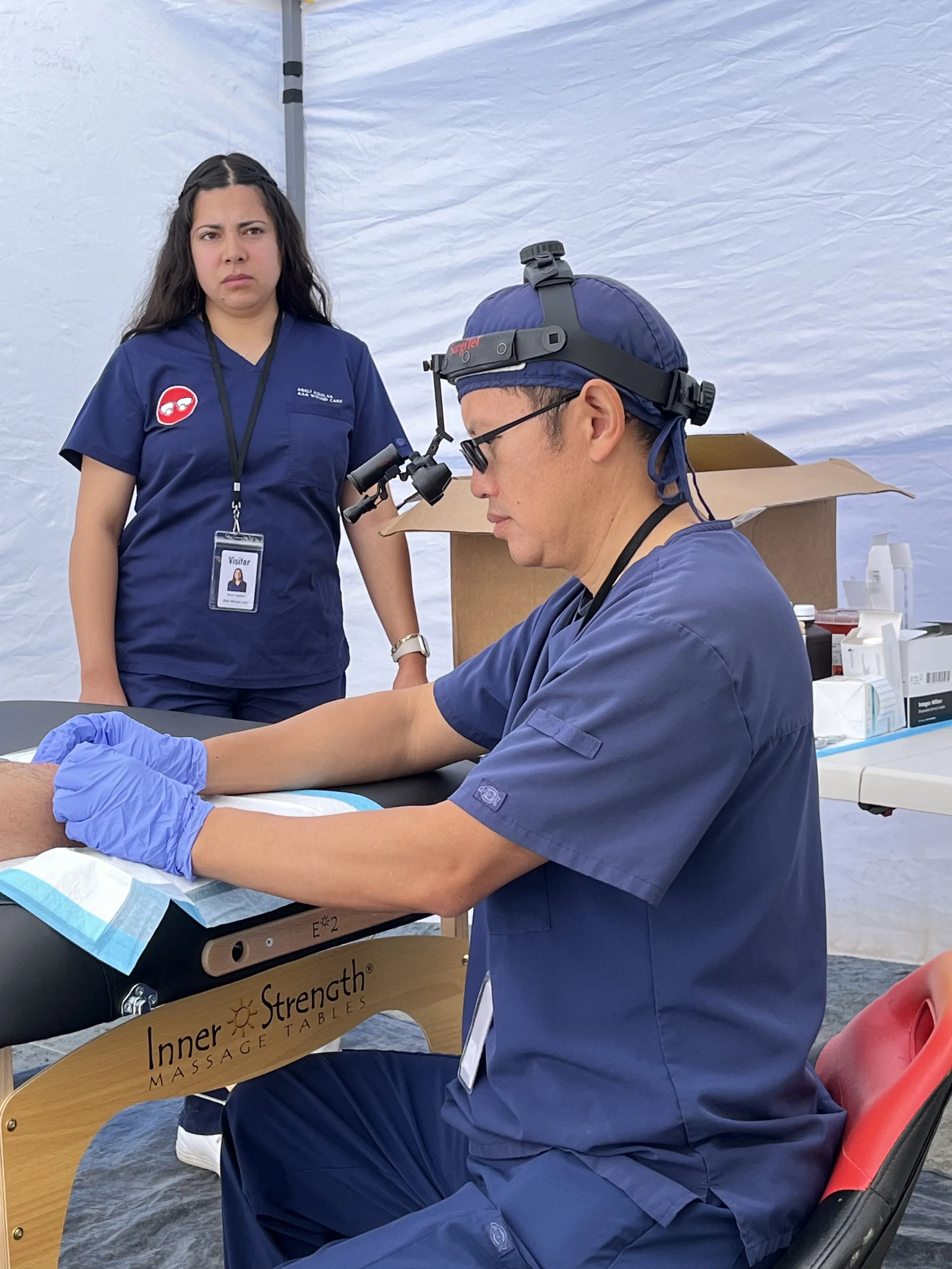 Medical professional in blue scrubs working on a patient's arm at an outdoor setup.