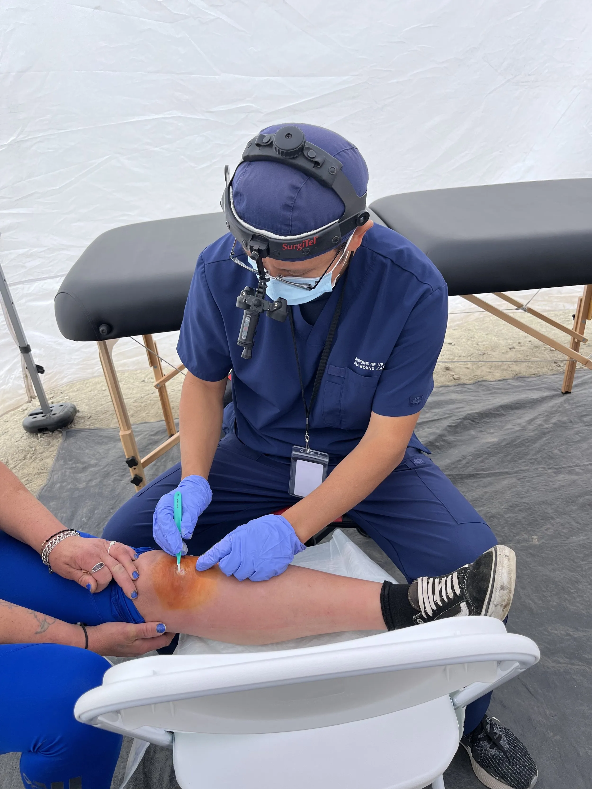 A medical professional in blue scrubs and surgical headgear performing a procedure on a patient's knee.