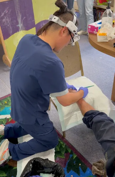 A medical professional wearing blue scrubs and a headlamp is kneeling and providing care to a patient's arm.