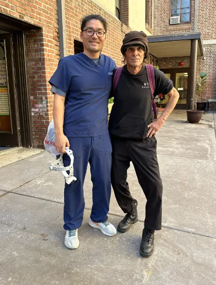 Two men posing outdoors, one in blue scrubs holding a medical device, the other in casual attire.