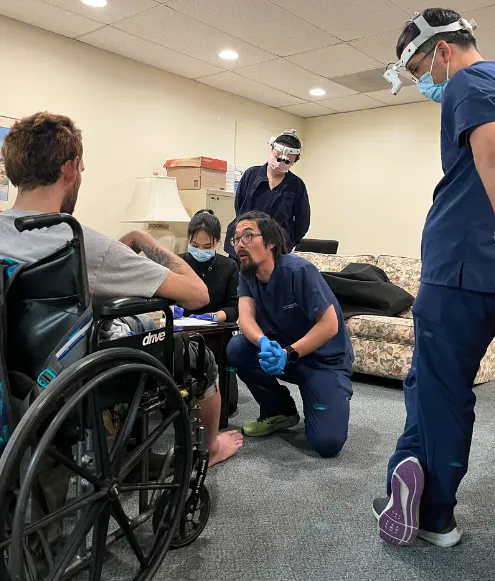 A team of healthcare professionals in scrubs, some wearing headgear and masks, are gathered around a person in a wheelchair.