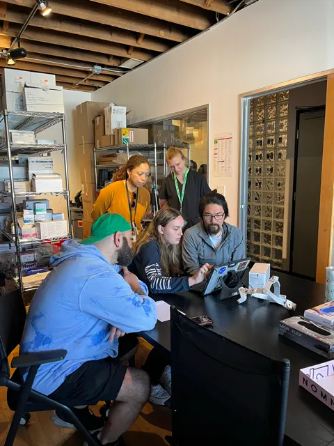 Five people in an office setting gathered around a table with a laptop.