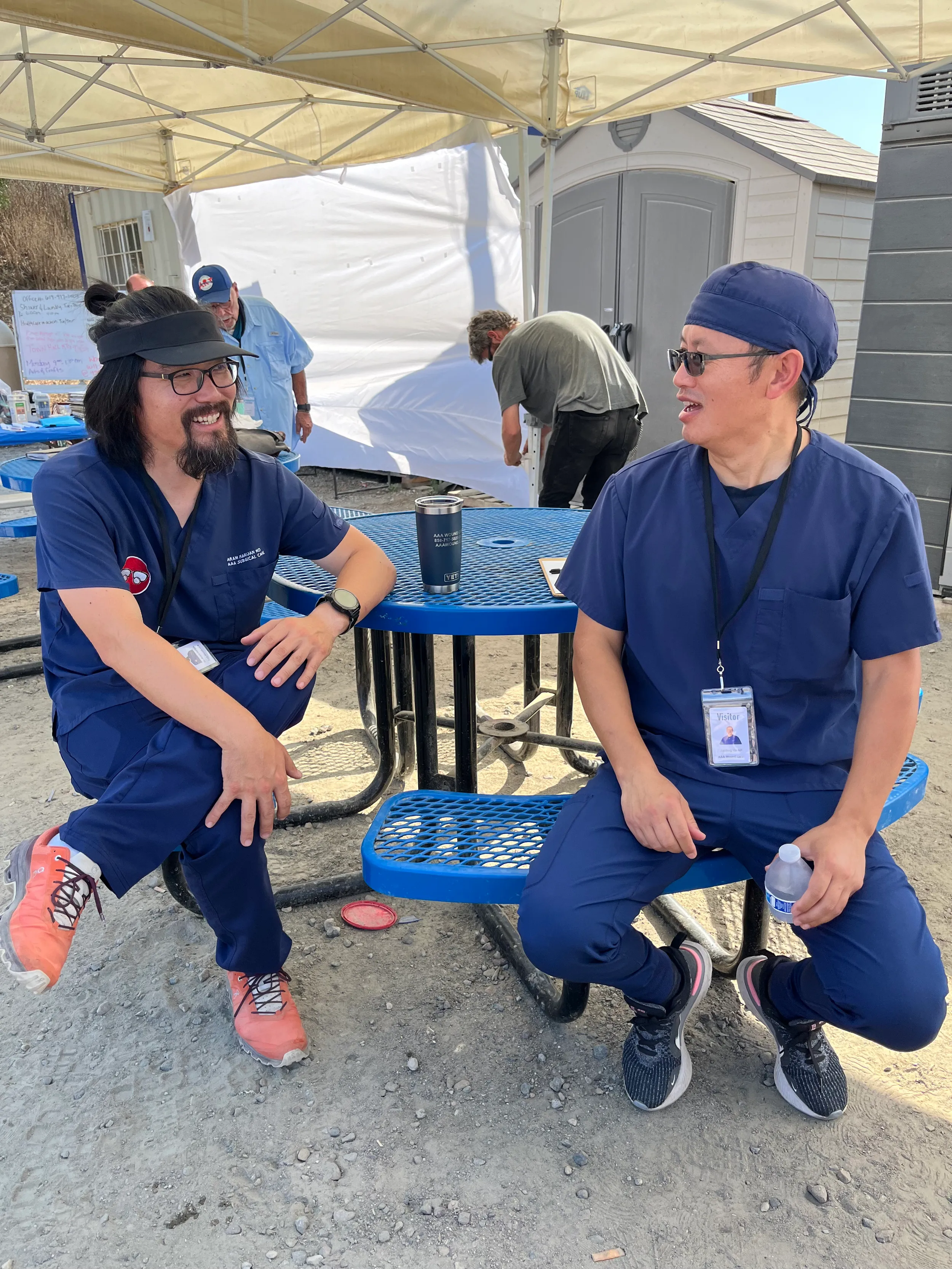 Two men in scrubs sitting and talking at a blue picnic table.